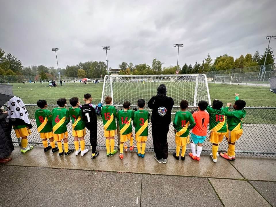 ACBC players in green and gold along the touchline at a rainy pitch